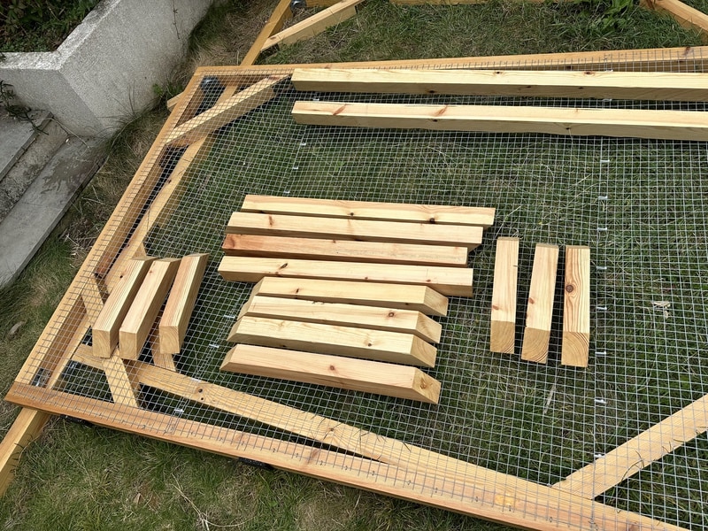 Two rectangles are lying in the grass. The top one has mesh over it, making it into a wall. More stabilising beams are resting on top. They look shiny since they've been varnished.