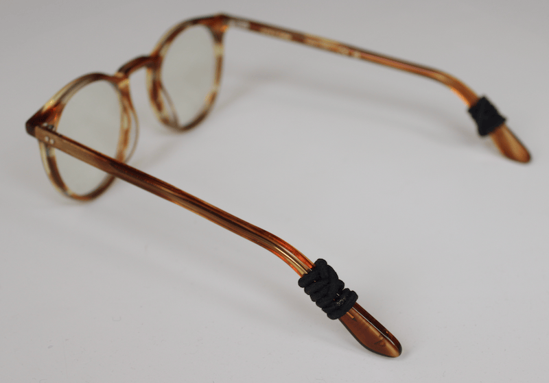 A pair of glasses with cream-colored plastic frames sitting on a white surface. Each temple has a black hair tie wrapped around it, just behind the part where it bends.
