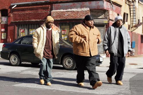 A screen capture from HBO’s The Wire. Three young, Black men are walking in the middle of a street. Behind them is a street corner, with a dilapidated, red brick building, with barred store-front windows. The men are wearing do-rags, a beanie, oversized coats, jeans, and Timberland boots. It’s a typical image of what White Americans think of, when they think of inner cities and urban decay.