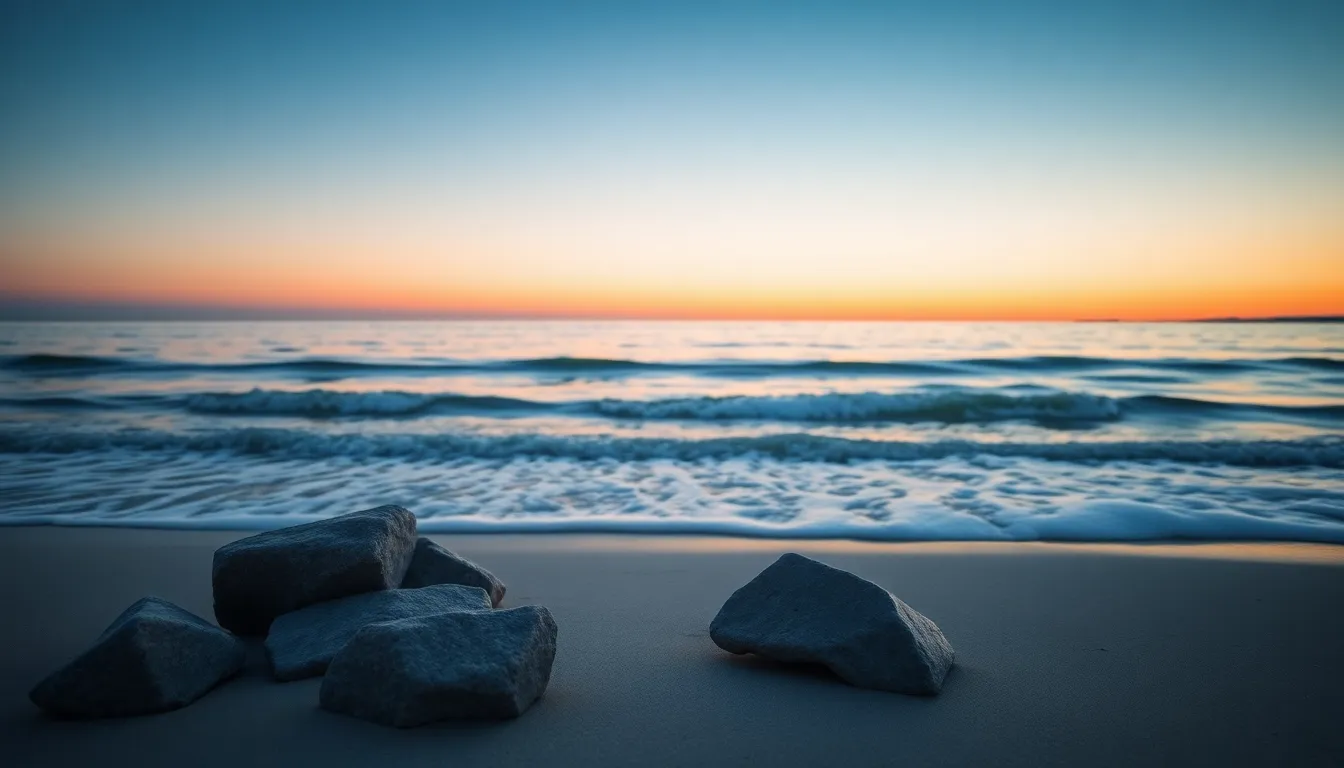 This serene beach scene captured at dawn showcases the beautiful transition of colors in the sky as day breaks. Soft pastel hues reflect gently on the water’s surface, creating a tranquil ambiance. A small group of boulders in the foreground adds depth, with their textures highlighted by the warm, diffused morning light. The composition and soft lighting evoke a sense of peace and harmony, perfect for those seeking a quiet retreat by the sea.