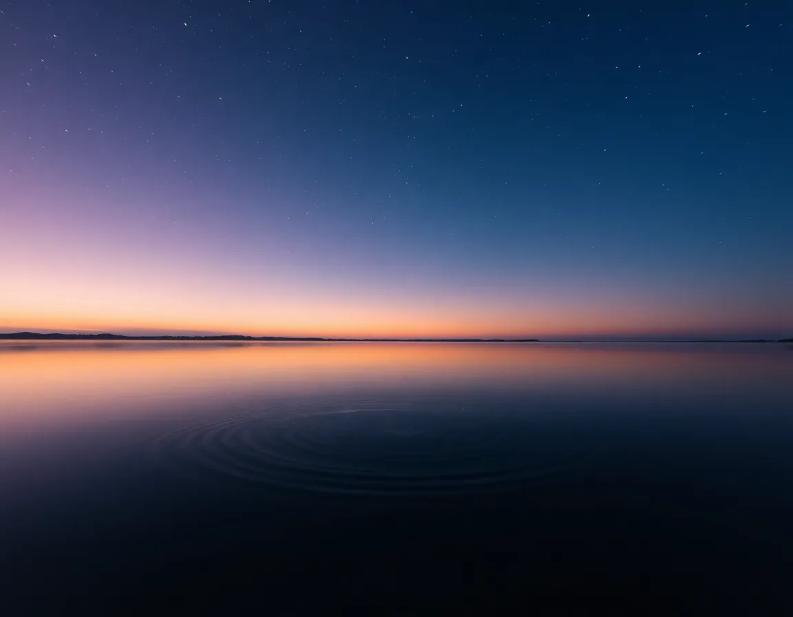 This serene photograph captures the reflective beauty of a star-filled sky mirrored in a tranquil lake at dawn. As the first light of day breaks, soft pinks and oranges blend with deep blue twilight, creating a mesmerizing pastel color palette. The shallow depth of field emphasizes the shimmering stars above while gentle ripples in the water enhance the peaceful ambiance. A centered composition beautifully showcases the symmetry between sky and reflection.