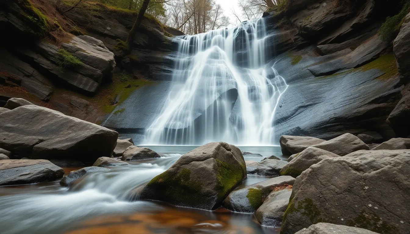 This serene waterfall scene is captured under soft overcast light, enveloping the landscape in natural muted tones. The calm flow of water contrasts beautifully with the textured moss-covered stones surrounding it. Using hyperfocal distance ensures that every detail from the rocks to the trees is in sharp focus, providing a rich visual experience. The composition centers on the waterfall, inviting viewers to immerse themselves in the tranquility of this natural setting.